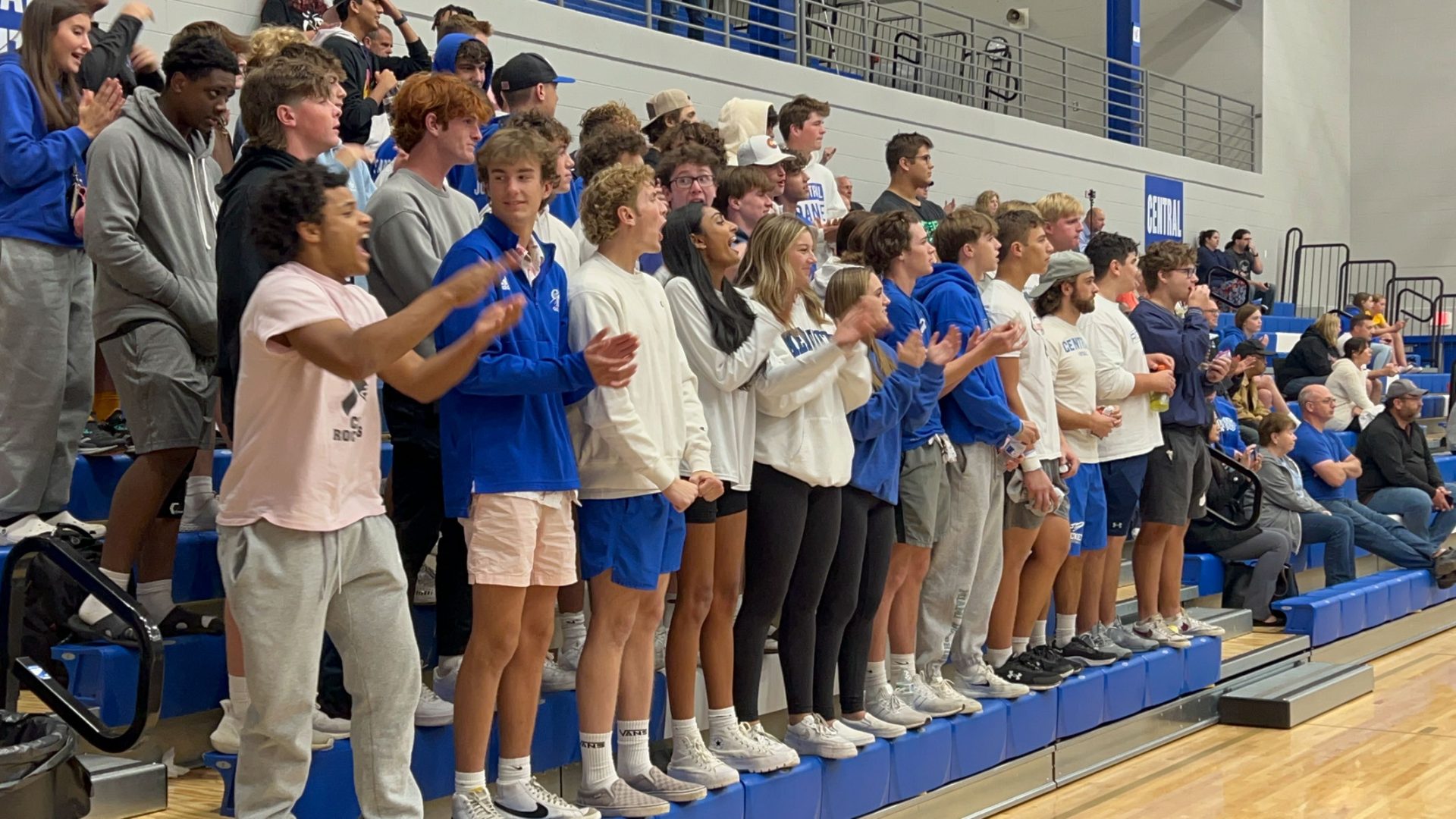 A group of high school students stands on gym bleachers, clapping and cheering.