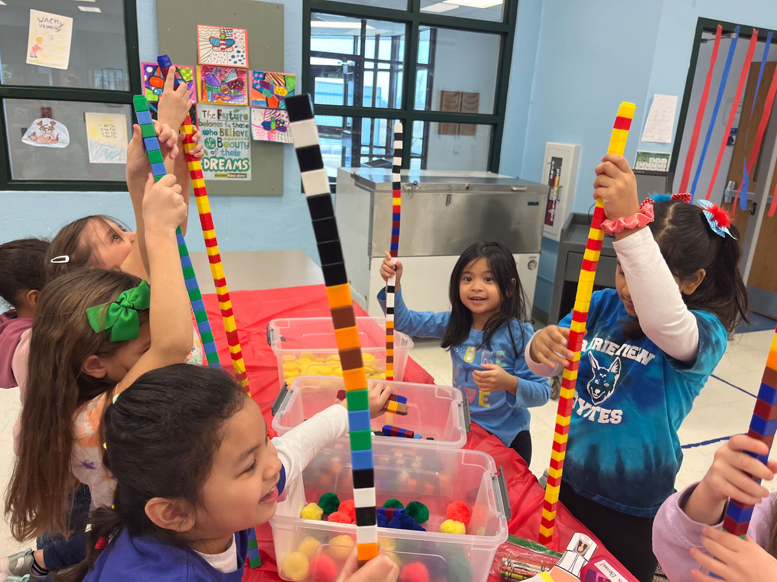 A group of grade school students in a classroom sitting around a table building towers made of interlocking plastic bricks.