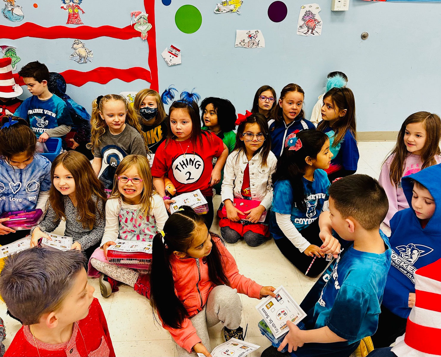 A group of kindergarten students holding pieces of paper sits together on a colorful classroom rug. The children are seated in a circle, surrounded by bright and cheerful classroom decorations. Some students are smiling.