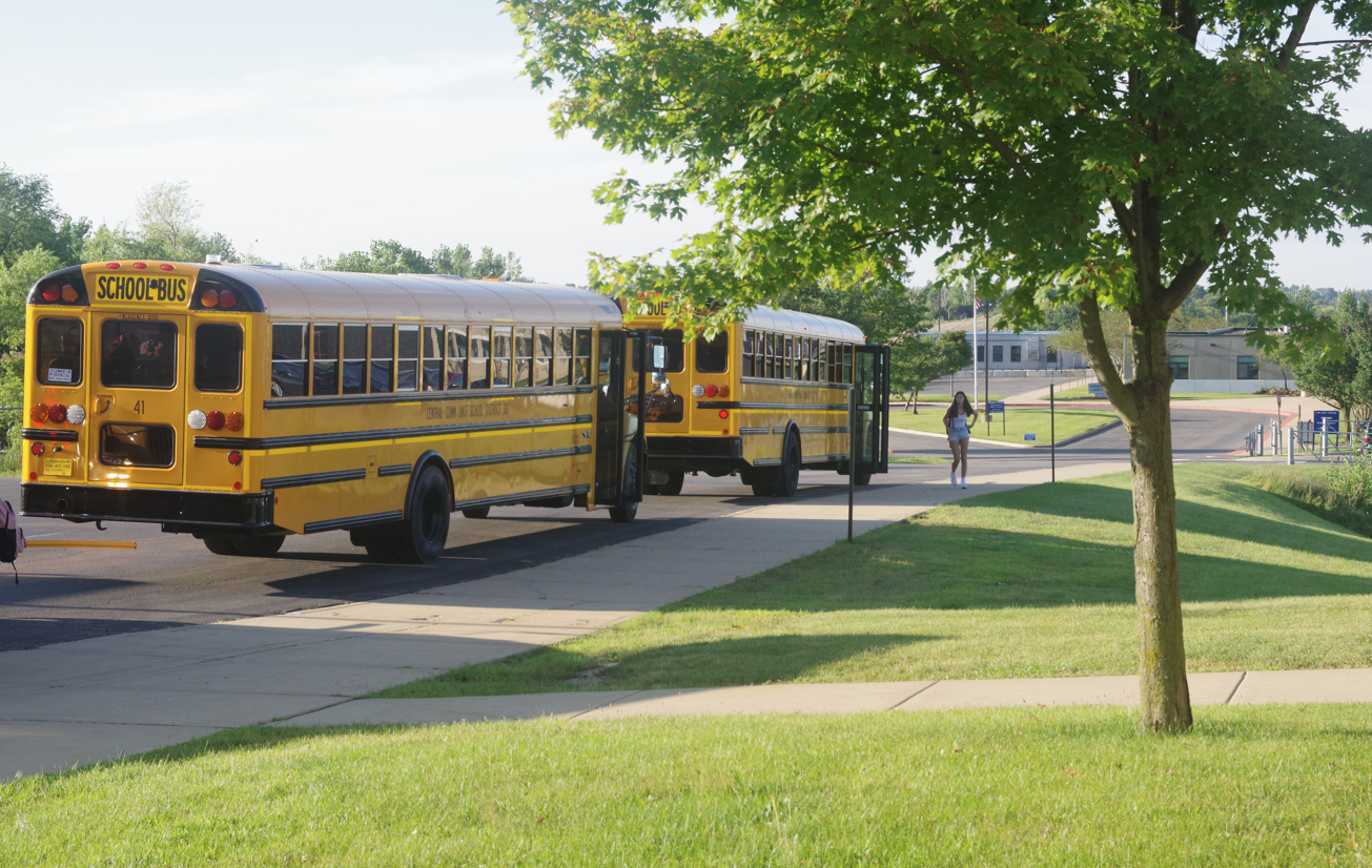 Two yellow school buses are parked alongside the curb of a grassy school yard, with a high school building visible in the background. A high school student walks on the sidewalk toward the buses.