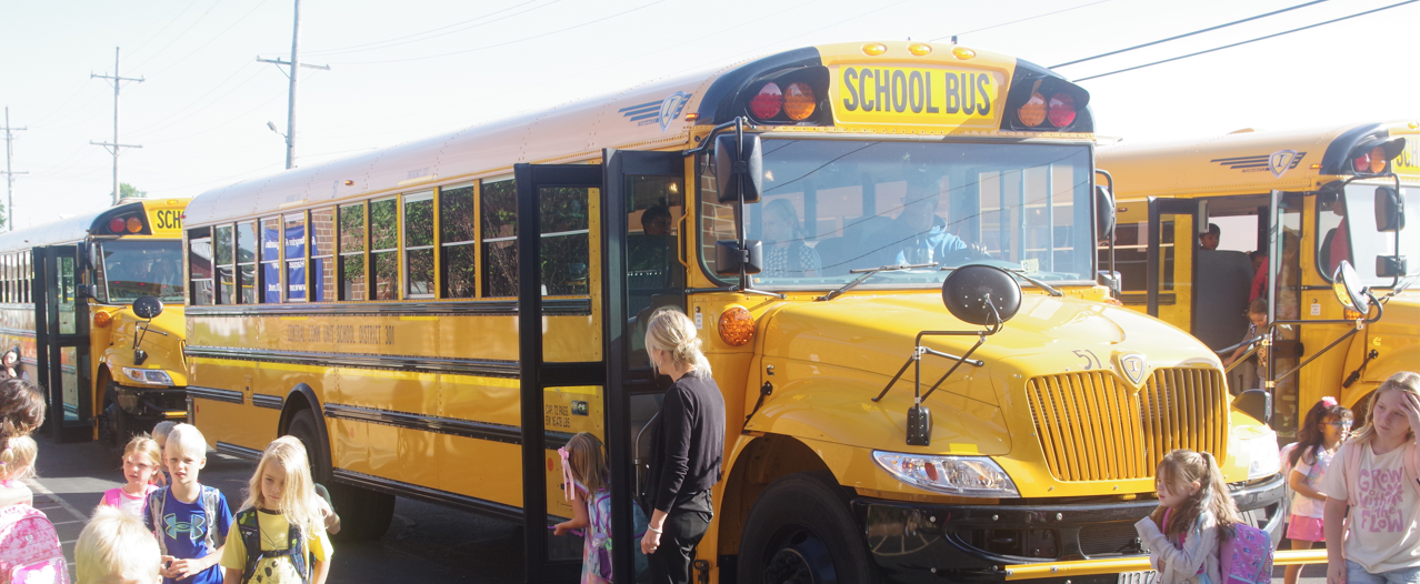 A group of yellow school buses is parked as young students get off the buses, with adults supervising the students' safe departure.