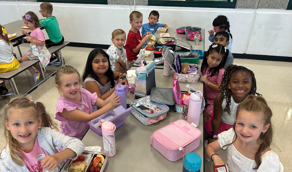 A group of grade school students sits at a lunch table in a school cafeteria, smiling at the camera. Their brightly-colored lunch boxes and thermoses are placed in front of them on the table.