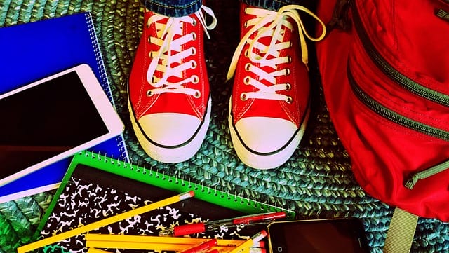 An older child's feet in bright red sneakers stand on a colorful classroom rug, surrounded by scattered school supplies, including notebook, smartphone, ruler, and pencils.