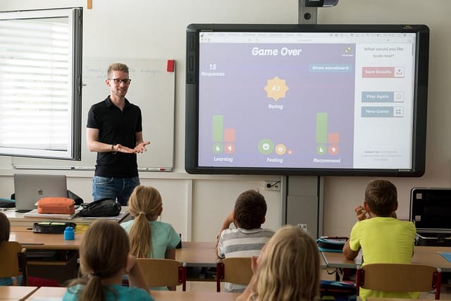 A teacher stands in front of a smartboard at the front of a classroom, with students sitting at desks visible from behind.